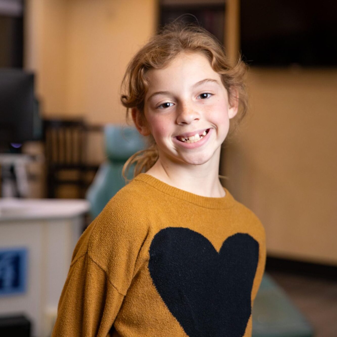Smiling girl with missing front teeth wearing a heart-patterned sweater in an orthodontic office setting, highlighting a welcoming environment for new patients.